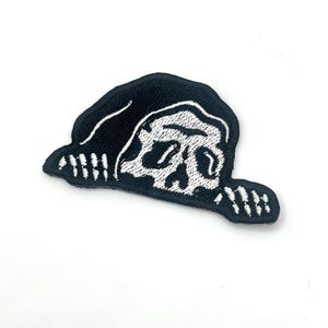 May include: A black and white embroidered patch featuring a skull wearing a hood. The skull is white with black detailing, and the hood is black. The patch has a stitched border and is likely for clothing or accessories.