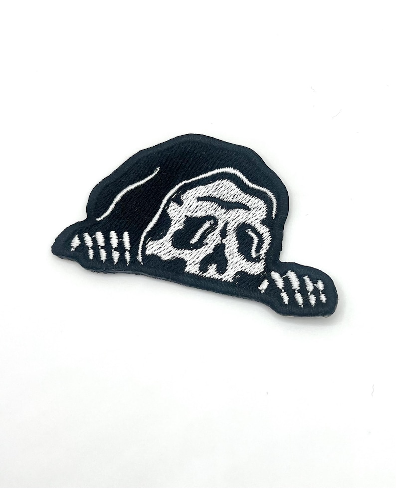 May include: A black and white embroidered patch featuring a skull wearing a hood. The skull is white with black detailing, and the hood is black. The patch has a stitched border and is likely for clothing or accessories.