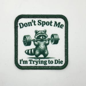 May include: Square patch with a white background and a green border. The patch features a raccoon lifting a barbell. Text on the patch reads "Don't Spot Me" above and "I'm Trying to Die" below the raccoon.