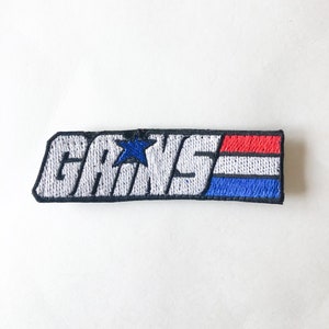 May include: A white embroidered patch with the word "G.I. JOE" in black lettering and a blue star. The patch has red, white, and blue stripes on the right side.