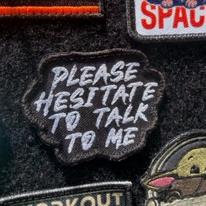 May include: A black embroidered patch with an irregular, cloud-like shape featuring the white text PLEASE HESITATE TO TALK TO ME in a bold, hand-lettered style. The patch has a dark border and is positioned among other various fabric patches.