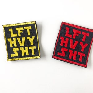 May include: Two black fabric patches with white stitching around the edges. The patches have the text "LFT HVY SHT" embroidered in yellow on one patch and red on the other.