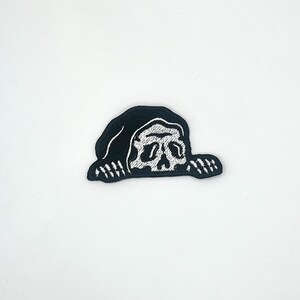May include: A black and white embroidered patch featuring a stylized skull design. The skull is white with detailed facial features, set against a black background. The patch has a unique, slightly irregular shape.
