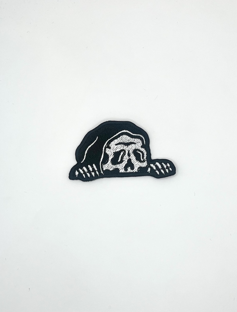 May include: A black and white embroidered patch featuring a stylized skull design. The skull is white with detailed facial features, set against a black background. The patch has a unique, slightly irregular shape.