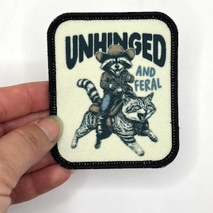 May include: A white patch with black stitching around the edges. The patch features a cartoon raccoon wearing a cowboy hat riding a cat. The text "Unhinged and Feral" is printed in black.