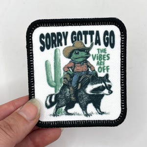 May include: A black and white patch featuring a green frog wearing a cowboy hat riding a raccoon. The text on the patch reads "Sorry Gotta Go The Vibes Are Off".