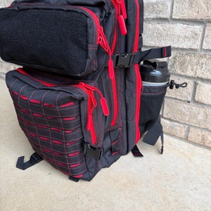 May include: Black backpack with red accents, zippers, and stitching. Features multiple compartments, a water bottle holder, and a black patch area. The bag has a tactical design with adjustable straps and buckles.