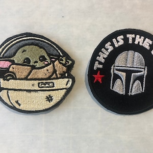 May include: Two embroidered patches, one featuring a cartoon character in a pod with the text "This is the way" and the other featuring a Mandalorian helmet with the text "This is the way".