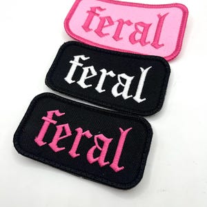 May include: Three embroidered patches with the word "feral". One patch is pink with pink text, another is black with white text, and the third is black with pink text. The patches have a rectangular shape with rounded corners.
