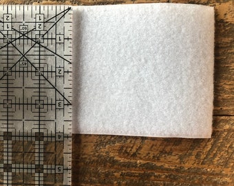 May include: A white, rectangular piece of fabric measuring approximately 3 inches by 4 inches. The fabric is placed on a wooden surface and is being measured with a ruler.
