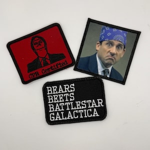 May include: Three embroidered patches. One red square patch features a black outline of a person and the text "CPR Certified". A black rectangular patch has the words "BEARS BEETS BATTLESTAR GALACTICA" in white. A square patch shows a person wearing a blue bandana.