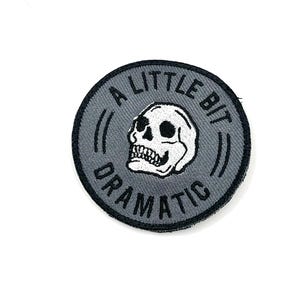 May include: Round, gray patch with the text "A LITTLE BIT DRAMATIC" encircling a white skull. The skull has black eye sockets and a detailed jawline. The patch has a black border and is likely for clothing or accessories.