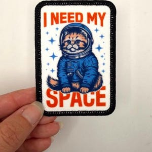 May include: A rectangular embroidered patch with a black border featuring a brown cat wearing a blue astronaut suit and helmet. The background is white with blue stars. The text I NEED MY SPACE is printed in orange capital letters.