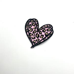 May include: A pink heart-shaped patch with a leopard print design. The patch has a black border and is set against a white background. The leopard print features tan spots on a pink base.