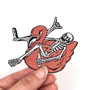 May include: Embroidered iron-on patch featuring a skeleton in a red inflatable tube with a drink in one hand and a shaka sign with the other.