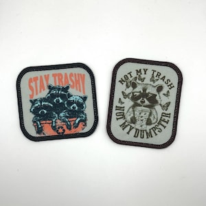 May include: Two square patches featuring raccoon designs. One says "Stay Trashy" with three raccoons in a bin. The other says "Not My Trash, My Dumpster" with a raccoon holding a cup, surrounded by butterflies.