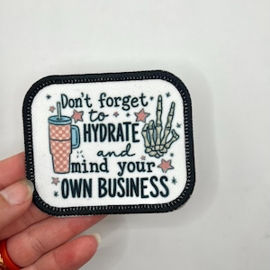 May include: A rectangular patch with a black border and the text "Don't forget to HYDRATE and mind your OWN BUSINESS." The patch features a pink and blue tumbler, a skeleton hand, and star accents.