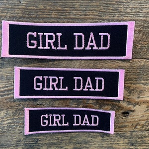 May include: Three black fabric patches with pink edging. Each patch says "GIRL DAD" in white lettering.