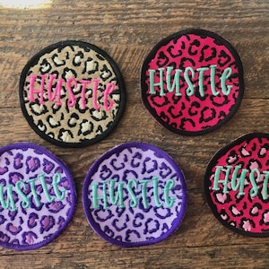 May include: Five round, embroidered patches with a leopard print background. Each patch has the word "Hustle" written in a light green font.