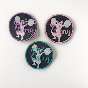 Puede incluir: Tres parches redondos negros con figuras bordadas blancas levantando pesas. Los parches tienen el texto "girl strong" en rosa, turquesa y morado.