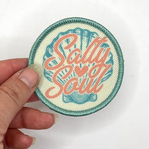 May include: Round patch with a light teal border and a cream-colored center. The patch features a seashell design in teal and the words "Salty Soul" in coral, with a small heart symbol.
