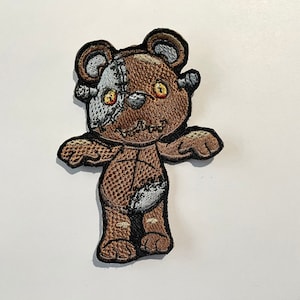 May include: Embroidered patch of a zombie teddy bear. The bear is brown with gray and white patches, stitched with black thread. It has yellow eyes and a stitched mouth, giving it a spooky appearance. The patch is on a white background.
