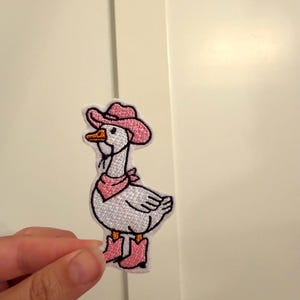 May include: An embroidered patch featuring a white goose wearing a pink cowboy hat, a pink bandana around its neck, and pink cowboy boots. The goose has an orange beak and black eyes, with black outlines defining the entire shape of the figure.