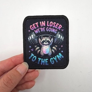 May include: A rectangular black patch with a stitched border featuring a graphic of a raccoon lifting a barbell. The text reads GET IN LOSER WE'RE GOING TO THE GYM in a colorful gradient font, surrounded by small star and heart shapes in pink and blue.
