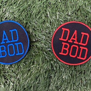 May include: Two round black fabric patches with white stitching around the edges. The patches have the text "DAD BOD" in blue and red embroidery.