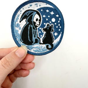 May include: Round embroidered patch featuring a black Grim Reaper figure and a black cat sitting on a crescent moon. The background is dark blue with white stars. The patch has a blue border and white stitching.