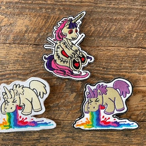 May include: Three embroidered iron-on patches featuring a unicorn design. The first patch shows a unicorn with a pink mane and tail, sitting down with its eyes closed. The second patch shows a unicorn with a rainbow coming out of its mouth. The third patch shows a unicorn with a purple mane and tail, also with a rainbow coming out of its mouth.