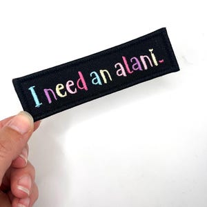 May include: Black rectangular patch with the embroidered text "I need an alani." The text is in a rainbow of colors, including blue, pink, yellow, and purple. The patch is held against a white background.