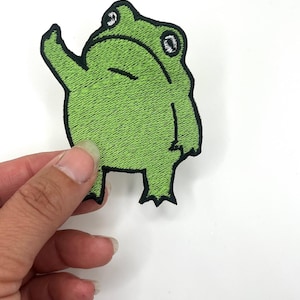 May include: Green embroidered frog patch with black outlines. The frog is standing on its hind legs with its front legs raised in a waving motion.