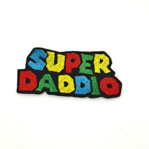 May include: Embroidered patch with the words "SUPER DADDIO" in colorful, block letters. The patch features a black border and a white background. The letters are blue, yellow, red, green, and blue.