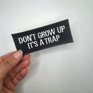 May include: Black embroidered patch with white text that reads "Don't grow up it's a trap".