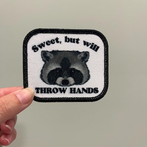 May include: Square patch with a black border and a raccoon graphic. The patch has the text "Sweet, but will THROW HANDS" in black, bold font. The raccoon is in shades of grey and black.