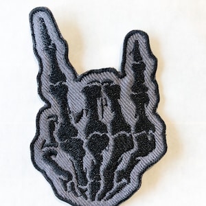 May include: A black and gray embroidered patch of a skeleton hand making the rock and roll hand gesture.
