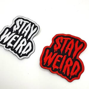 May include: Two embroidered patches with the words "STAY WEIRD". One patch is black with white lettering and the other is red with black lettering. Both patches have a white background.