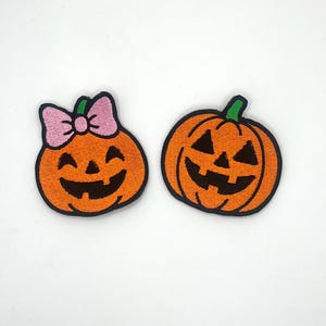 May include: Two embroidered pumpkin patches. One orange pumpkin has a green stem and a smiling face with black outlines. The other pumpkin has a pink bow, green stem, and a smiling face with black outlines. Both patches have black outlines.