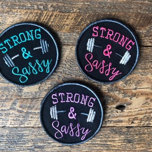 Peut inclure: Trois patchs ronds noirs à repasser avec des broderies blanches et grises. Les patchs disent "Strong & Sassy" avec un graphique de barre. Les patchs ont des broderies de différentes couleurs : turquoise, rose et violet.