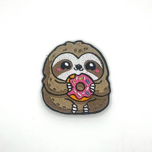 May include: A brown sloth patch with a cute design, holding a pink donut with sprinkles. The sloth has large eyes and rosy cheeks. The patch is on a white background.