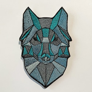 Peut inclure: Un patch brodé représentant une tête de loup géométrique dans des tons de bleu et de gris. Le visage du loup est stylisé avec un motif géométrique.