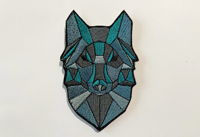 May include: A geometric wolf head embroidered patch in shades of blue and gray. The wolf's face is stylized with a geometric pattern.
