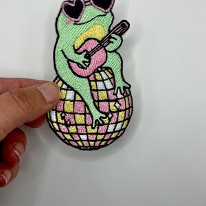 May include: Green frog wearing pink heart-shaped sunglasses and playing a pink guitar while sitting on a pink and yellow disco ball.
