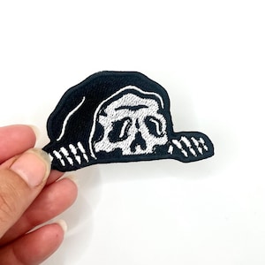 May include: Embroidered patch featuring a skull wearing a hat. The patch is black with white stitching, depicting the skull's features and hat details. The patch is held against a white background.