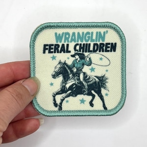 May include: A light green square patch with a white background and a black and blue illustration of a cowgirl riding a horse. The text "WRANGLIN' FERAL CHILDREN" is printed in black letters on the patch.