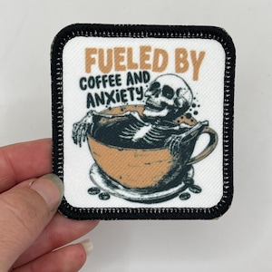 May include: A white square patch with black stitching around the edges. The patch features a cartoon skeleton submerged in a cup of coffee. The text "Fueled by Coffee and Anxiety" is printed above the skeleton.