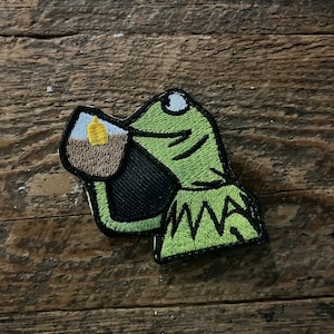 May include: Green felt patch of a frog drinking from a brown mug with a yellow lid. The frog has a white eye and a black outline.