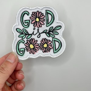 May include: White embroidered patch with green and pink flowers and the text "God is Good" in black.