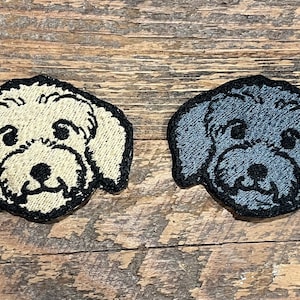 May include: Two embroidered dog patches, one white and one gray, with black outlines. The patches are on a wooden surface.